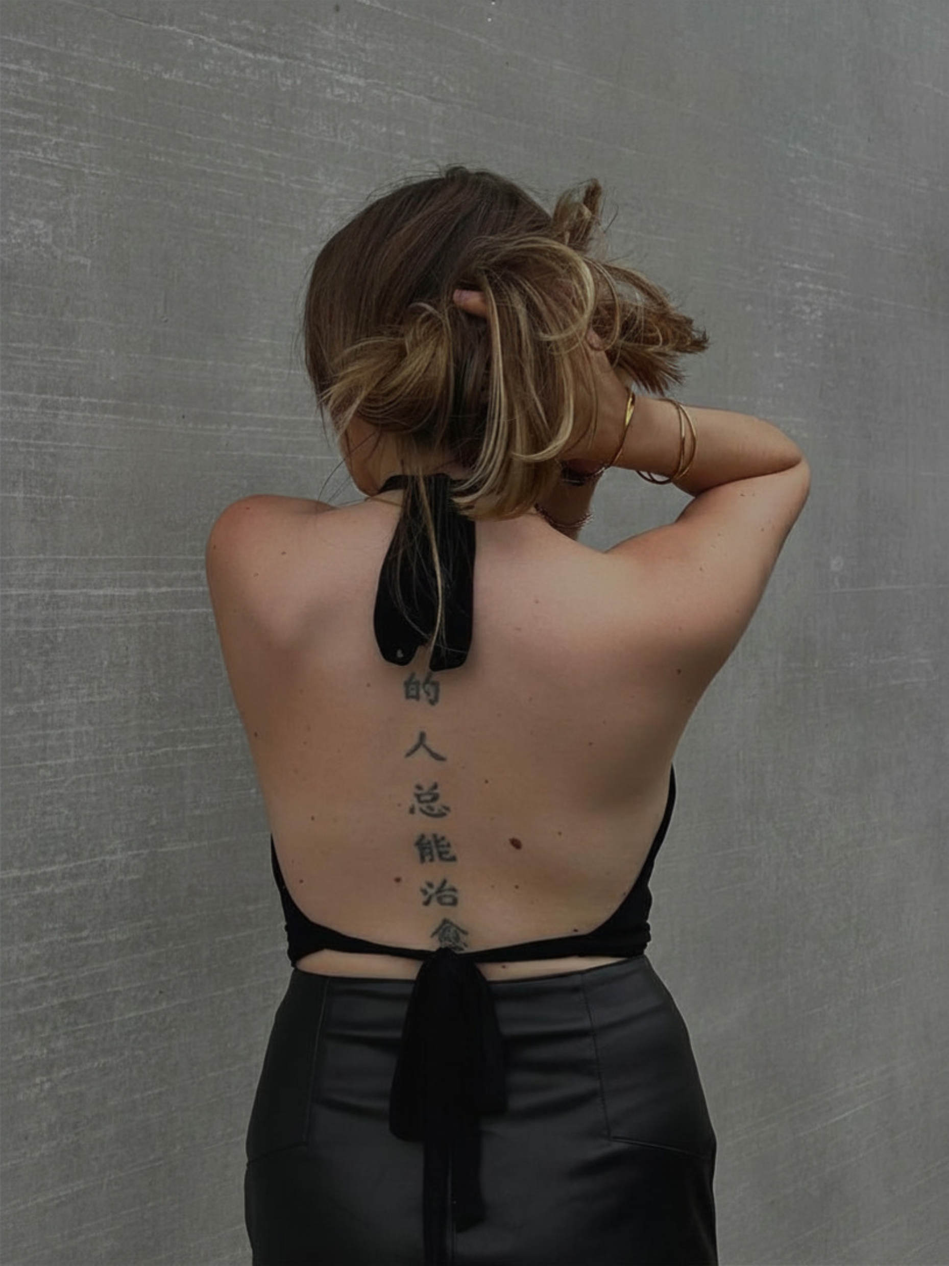 Portrait de dos d'une femme relevant ses cheveux pour dévoiler le grand dos nu du haut ajustable noir de la marque Bonjour Leoni, avec ses lanières nouées délicatement autour du cou et de la taille.